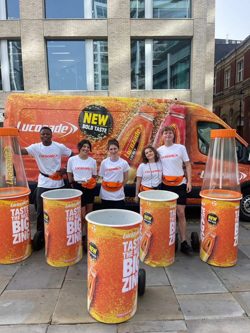 Group of individuals poses in front of a Lucozade van, with large bottles of Lucozade displayed nearby.